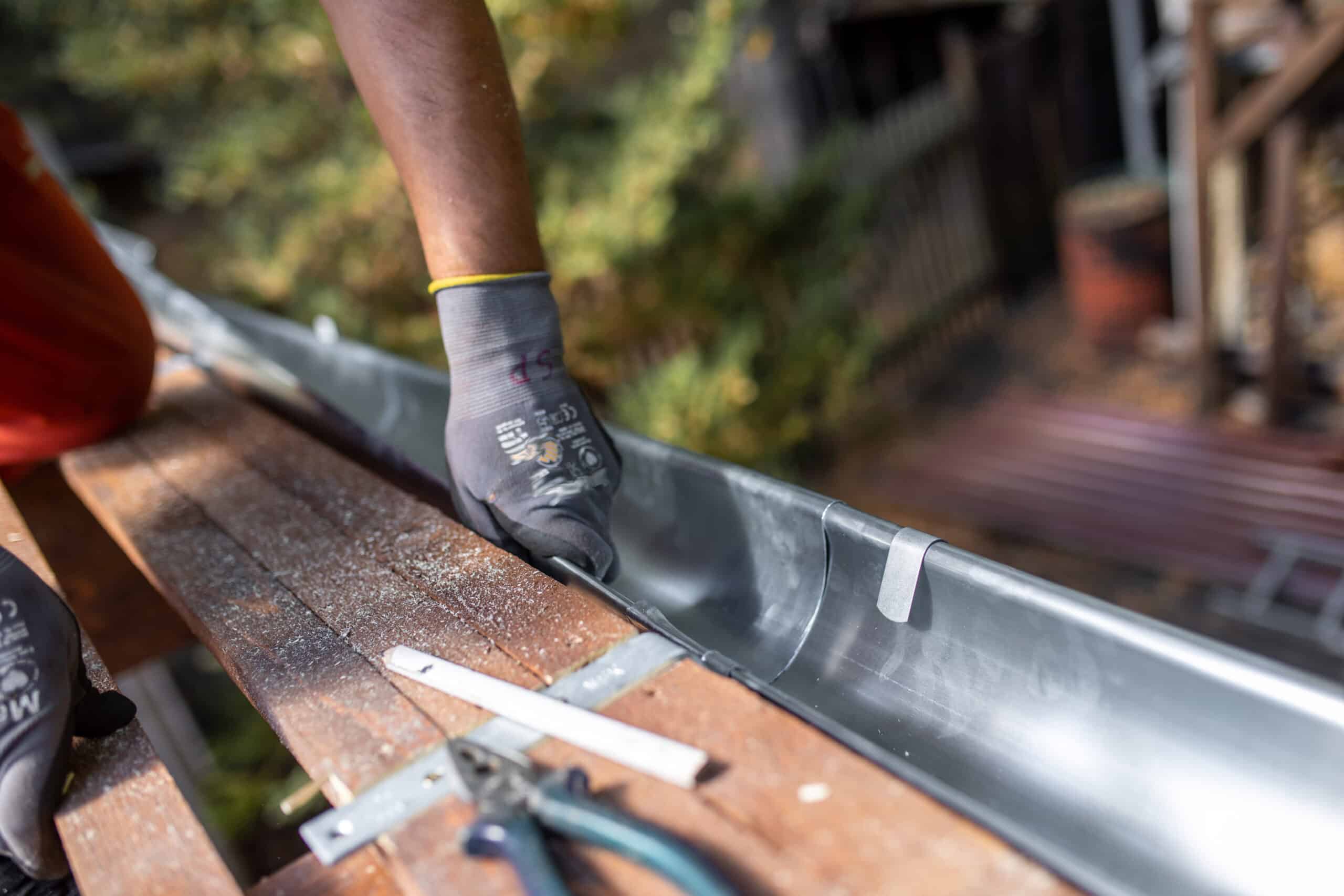 Gant de travail gris ajustant une gouttière en plomb ou zinc posée sur des planches de bois, avec des outils à proximité.