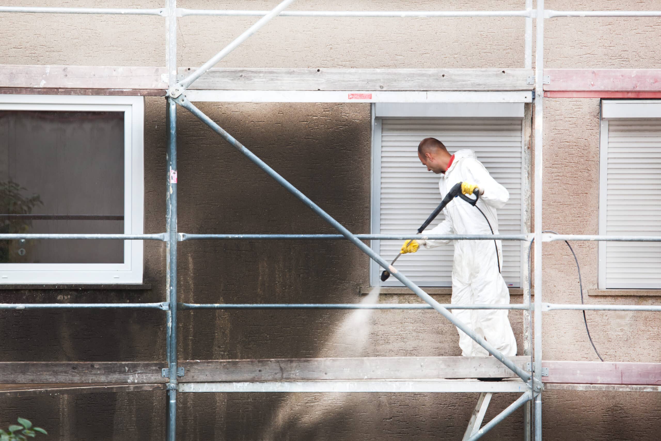 Ouvrier en combinaison blanche et gants jaunes nettoie une façade sale avec un nettoyeur haute pression depuis un échafaudage.